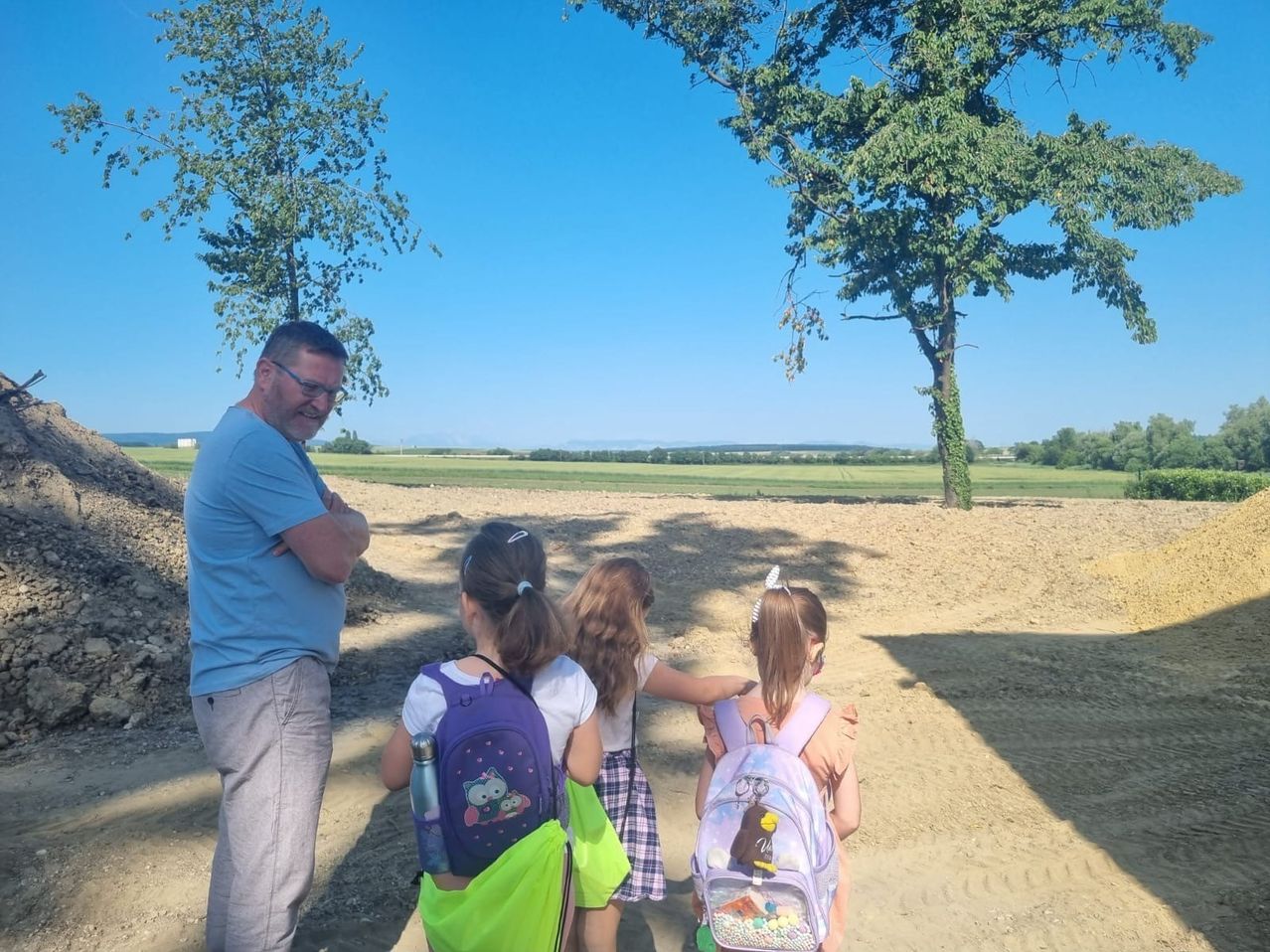 Ein Mann mit Brille steht auf einem Feld mit drei jungen Mädchen, alle tragen Rucksäcke, unter einem Baum.