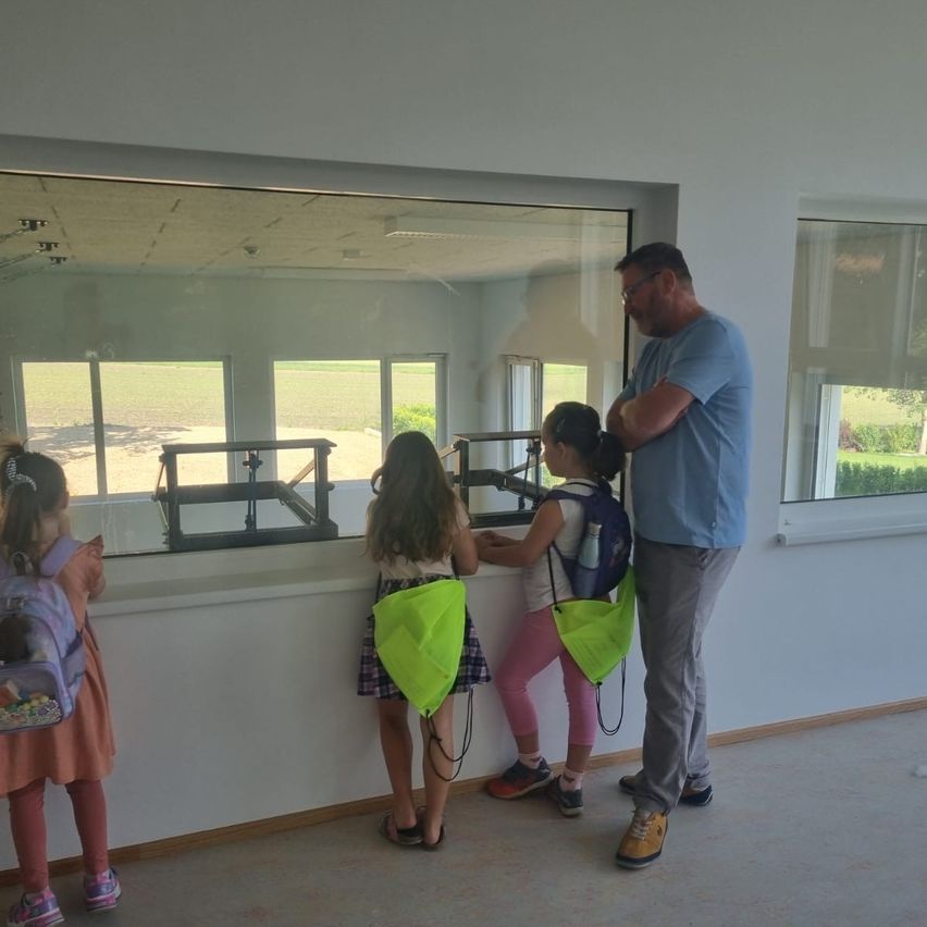 Drei junge Mädchen mit Rucksäcken stehen in einem Raum und schauen durch ein großes Fenster auf ein Feld. Ein Mann steht hinter ihnen und schaut ebenfalls aus dem Fenster.