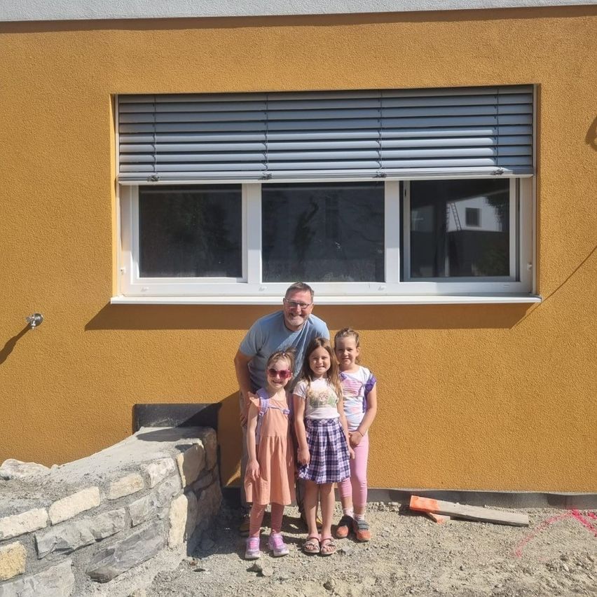 Ein Mann und drei junge Mädchen stehen vor einem gelben Gebäude mit einem weißen Fenster. Die Mädchen tragen Kleider und Sandalen. Der Mann trägt eine Brille und ein blaues Hemd.