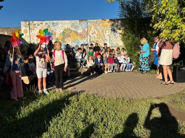 Children and adults gather in an outdoor area with painted walls and grassy ground. Some hold colorful pinwheels. An adult woman stands on a circular path, possibly speaking to the children.