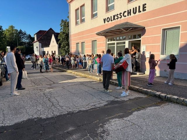 A crowd of people are standing outside a pink building with the sign 'VOLKSSCHULE'. Some are waiting in line while others are talking. There are trees and a house in the background.