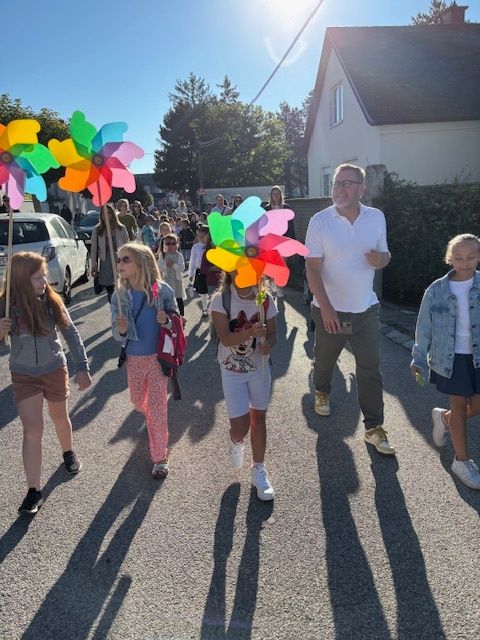 A group of children and adults walk down a street with colorful pinwheels on a sunny day. A man and a girl walk in the front.