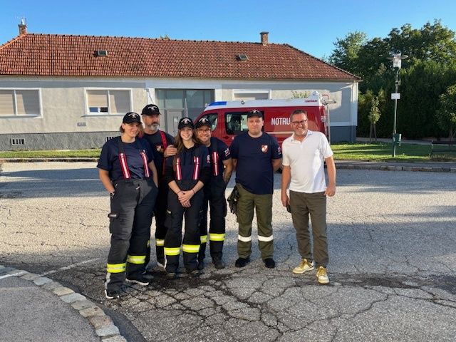 A group of firefighters stands in front of a fire truck, smiling and posing for a photo. Behind them is a building with a tiled roof.
