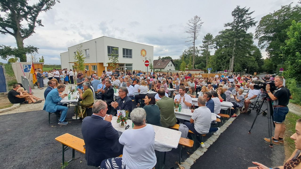Many people are gathered around tables in an outdoor area. Some are clapping. Trees and a building are in the background.