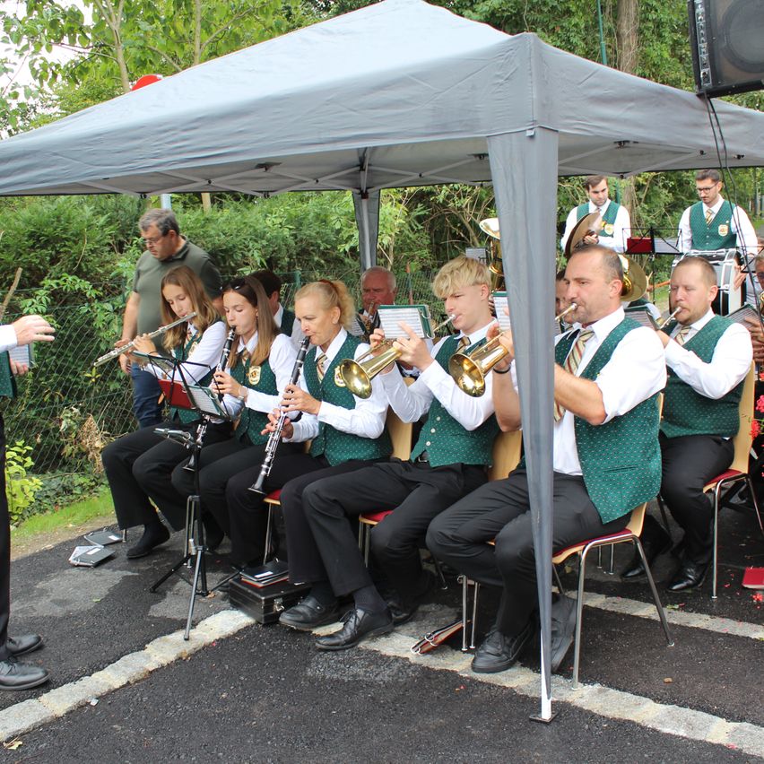 A band under a tent plays instruments, including trumpets and clarinets. Several musicians sit on chairs while others stand behind them. The setting is outdoors with trees and greenery in the background.
