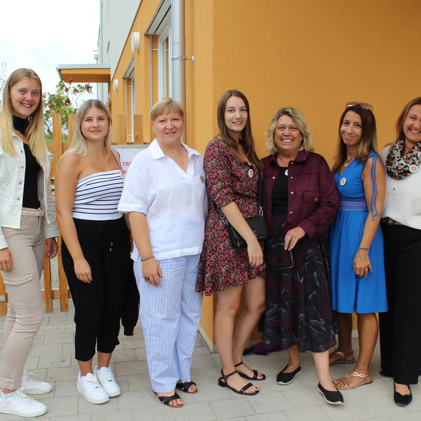 A group of women stand together outside a yellow building. They are smiling and appear to be posing for a photograph. They wear various outfits and accessories.