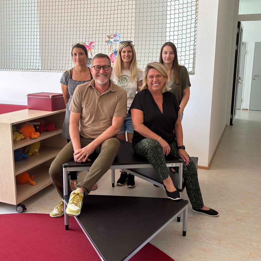 Fünf Personen, darunter ein Mann, sitzen auf Bänken in einem Raum mit rotem Teppich. Sie lächeln und posieren für ein Foto. Ein Holzschrank mit Spielzeug steht links.