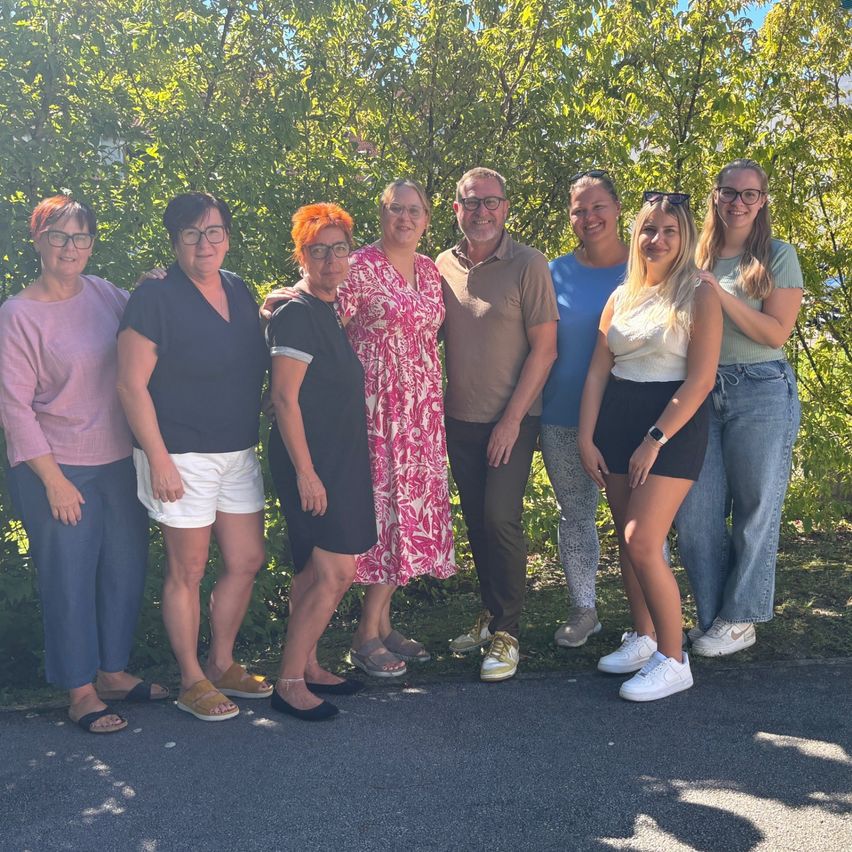 Eine Gruppe von acht Erwachsenen, darunter Männer und Frauen, posiert für ein Foto vor grünen Bäumen und einem Gebäude an einem sonnigen Tag.