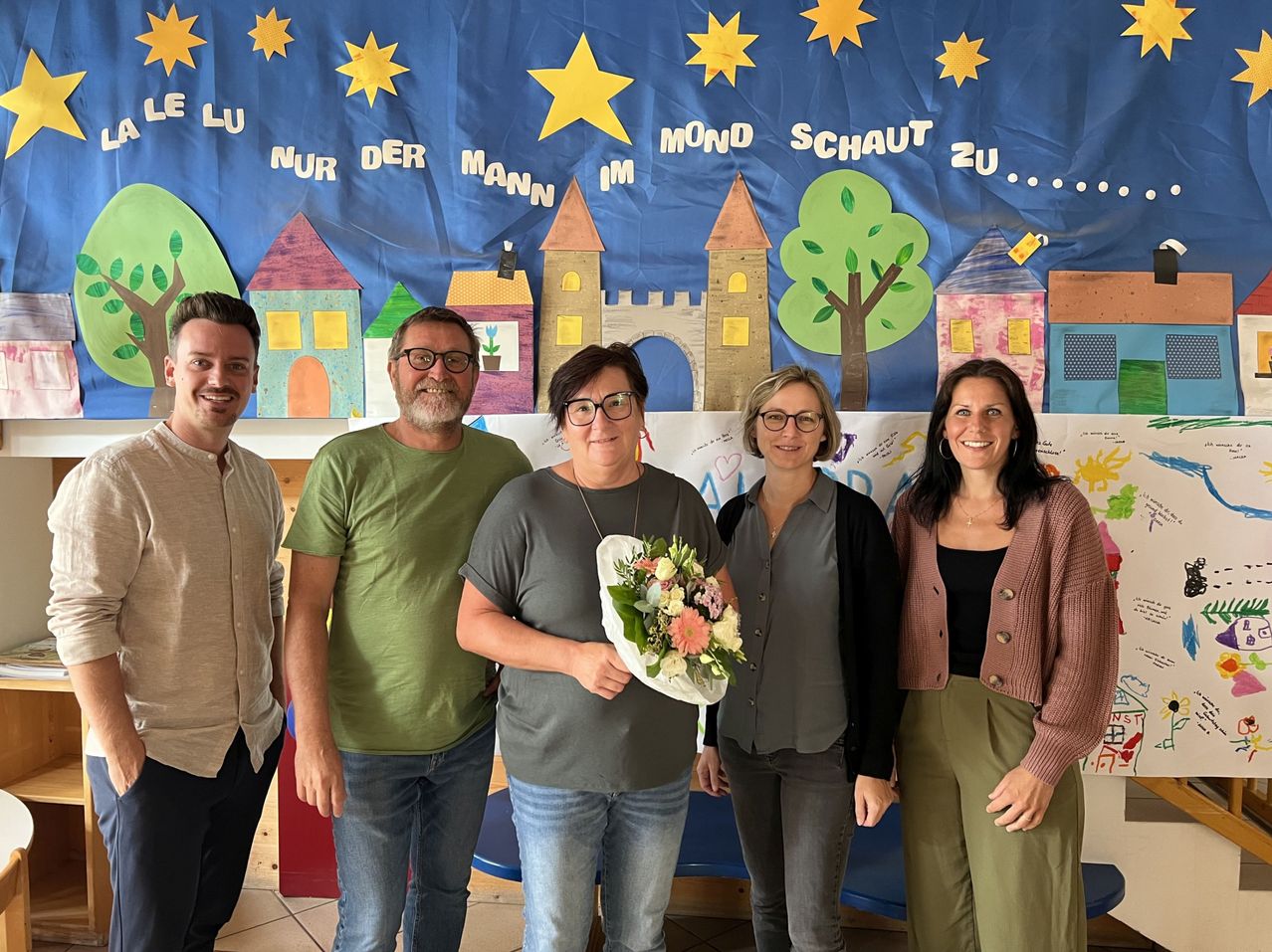 Fünf Personen posieren vor einem Hintergrund mit einem Banner, auf dem eine Zeichnung einer Stadt mit Häusern und einer Burg sowie einem Baum und Sternen abgebildet ist. Die Frau in der Mitte hält einen Blumenstrauß.