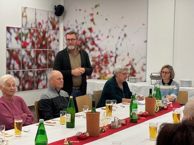 Eine Gruppe von Menschen sitzt an einem langen Tisch mit Getränken und Essen. Ein Mann steht hinter ihnen und sieht nachdenklich aus. Hinter dem Tisch befindet sich ein Wandbild mit Herbstblättern.