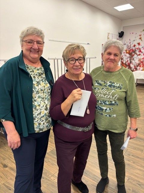 Drei Frauen stehen lächelnd in einem Raum. Eine hält ein Papier, eine andere eine Medaille. Sie tragen Freizeitkleidung und stehen auf einem Holzboden.