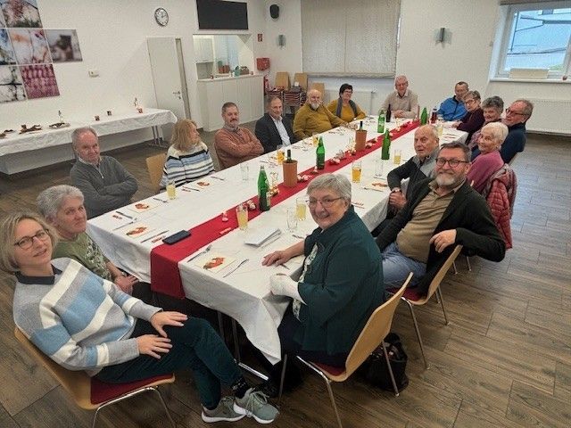 Eine Gruppe von Menschen sitzt um einen langen Tisch in einem Raum und posiert für ein Foto. Flaschen, Gläser und Teller sind auf dem Tisch.