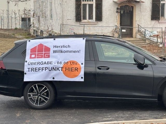 Ein schwarzer Wagen mit einem Banner, auf dem 'Herzlich willkommen! Übergabe - 08:00 Uhr Treffpunkt hier' steht, ist vor einem Haus mit Treppen geparkt.