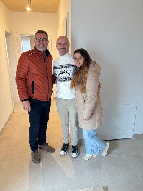 Drei Menschen stehen zusammen in einem Flur. Ein Mann mit Brille und einer roten Jacke, ein kahlköpfiger Mann in einem weißen Rollkragenpullover und eine Frau in einer beigen Jacke. Sie scheinen für ein Foto zu lächeln.