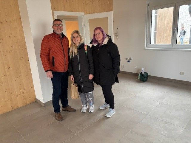 Drei Personen stehen in einem leeren Raum und lächeln für ein Foto. Der Raum hat geflieste Böden, weiße Wände und eine Holzwand. Ein Fenster befindet sich rechts, und eine Person ist draußen.