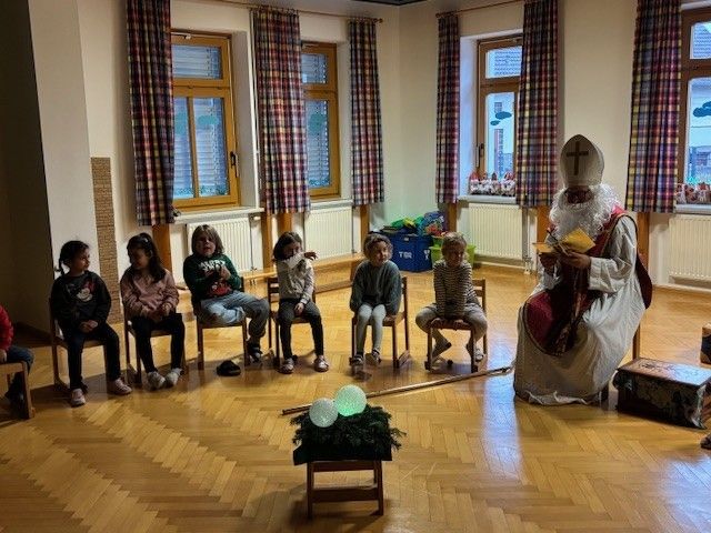 Mehrere Kinder sitzen auf Stühlen und lauschen einem Mann, der als Weihnachtsmann verkleidet ist. Er hält ein Buch in der Hand und liest vor.