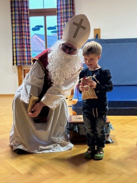 Eine als Papst verkleidete Person gibt einem Kind in einem Raum mit Holzboden und einem Fenster mit Vorhängen ein Geschenk.