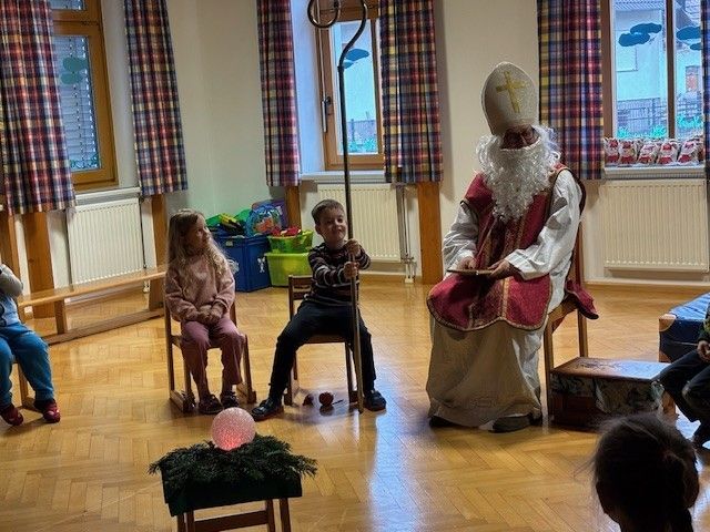 Eine als Bischof verkleidete Person liest Kindern, die auf Stühlen sitzen, ein Buch vor. Eine rosa Lampe steht auf einem kleinen Tisch vor ihnen.