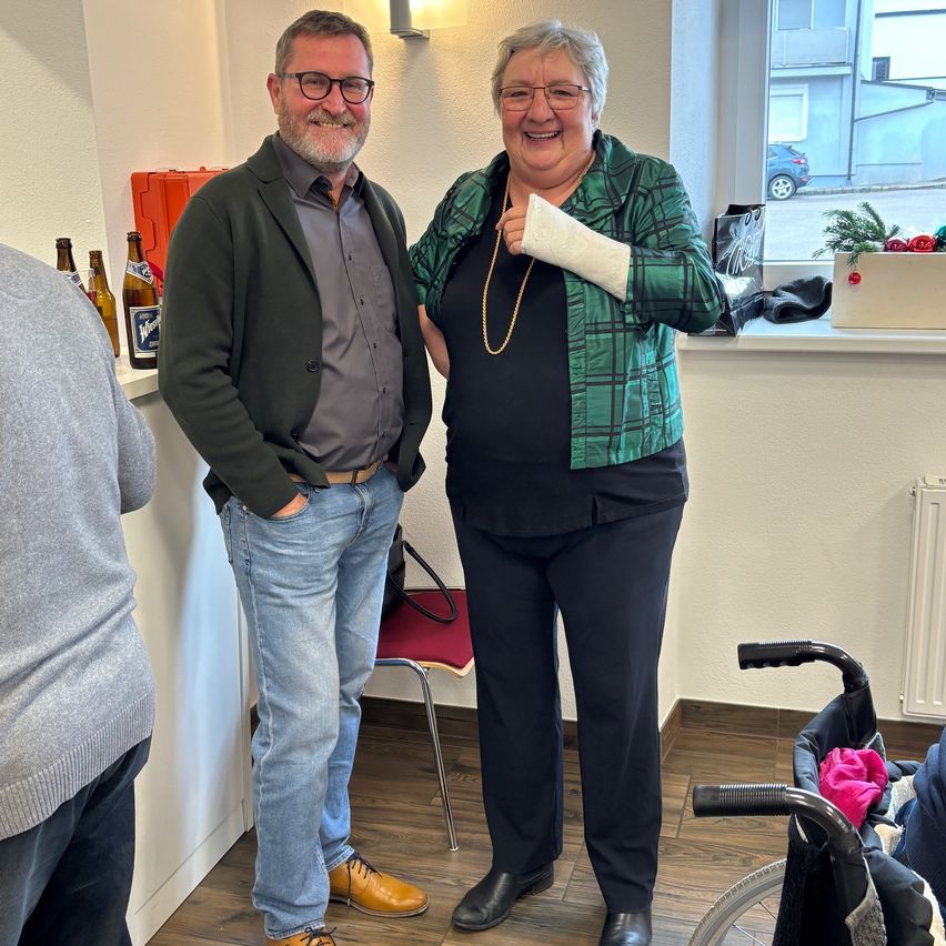 Ein Mann und eine Frau stehen lächelnd nebeneinander in einem Raum. Der Mann trägt ein graues Hemd und Jeans, während die Frau eine grüne Jacke und schwarze Hosen hat. Hinter ihnen ist eine weiße Wand mit einem Regal, das Flaschen und eine rote Box hält. Ein Stuhl steht in der Nähe des Fensters mit Blick auf ein Auto.