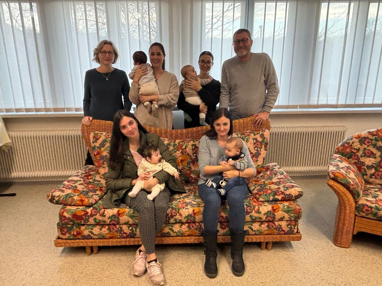 Eine Familie ist in einem Wohnzimmer versammelt. Zwei Frauen sitzen auf einem Blumensofa, jede hält ein Baby. Hinter ihnen stehen drei Personen, die ebenfalls Babys halten. Der Raum hat große Fenster mit weißen Vorhängen.