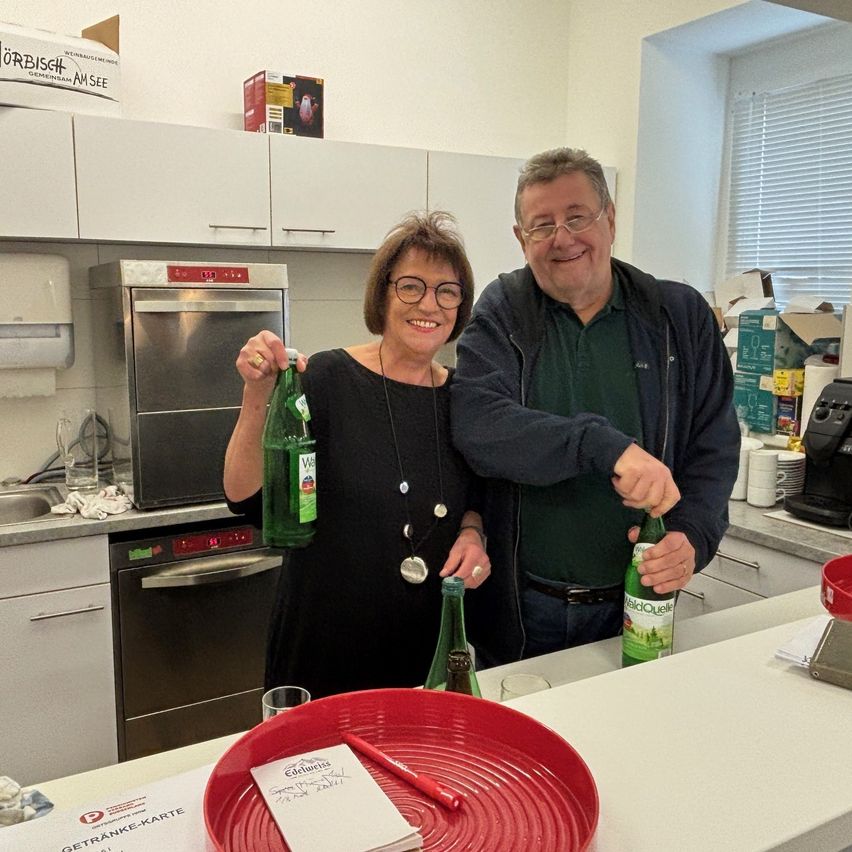 Ein Mann und eine Frau stehen in einer Küche und lächeln. Die Frau hält eine grüne Flasche und der Mann auch. Sie sind von Küchenutensilien umgeben, einschließlich eines Waschbeckens und einer Spülmaschine.