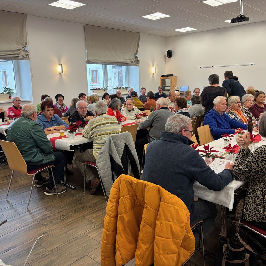 Eine Gruppe von Menschen ist in einem Raum versammelt und sitzt an langen Tischen, die mit weißen Tüchern bedeckt sind. Sie sind von dekorativen Gegenständen und einer Projektionsleinwand umgeben. Einige Personen tragen Brillen und Jacken, und der Raum ist von Lichtern beleuchtet.