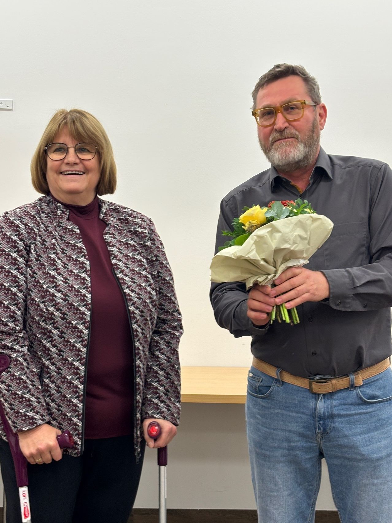 Eine Frau in einer gemusterten Jacke steht neben einem Mann mit Brille und Gürtel, beide lächeln, wobei der Mann einen Blumenstrauß hält. Sie stehen vor einer weißen Wand mit einem Holztisch dahinter.