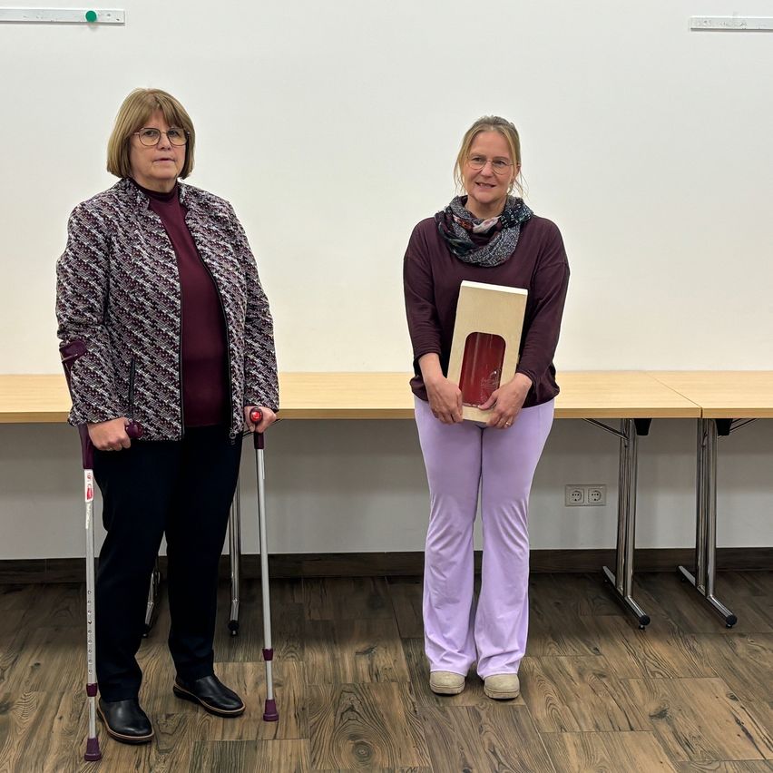 Zwei Frauen stehen in einem Raum. Eine hält eine Box und die andere eine Krücke. Sie lächeln beide. Der Raum hat eine weiße Wand und einen Holzboden. Hinter ihnen befinden sich Tische und eine Steckdose.