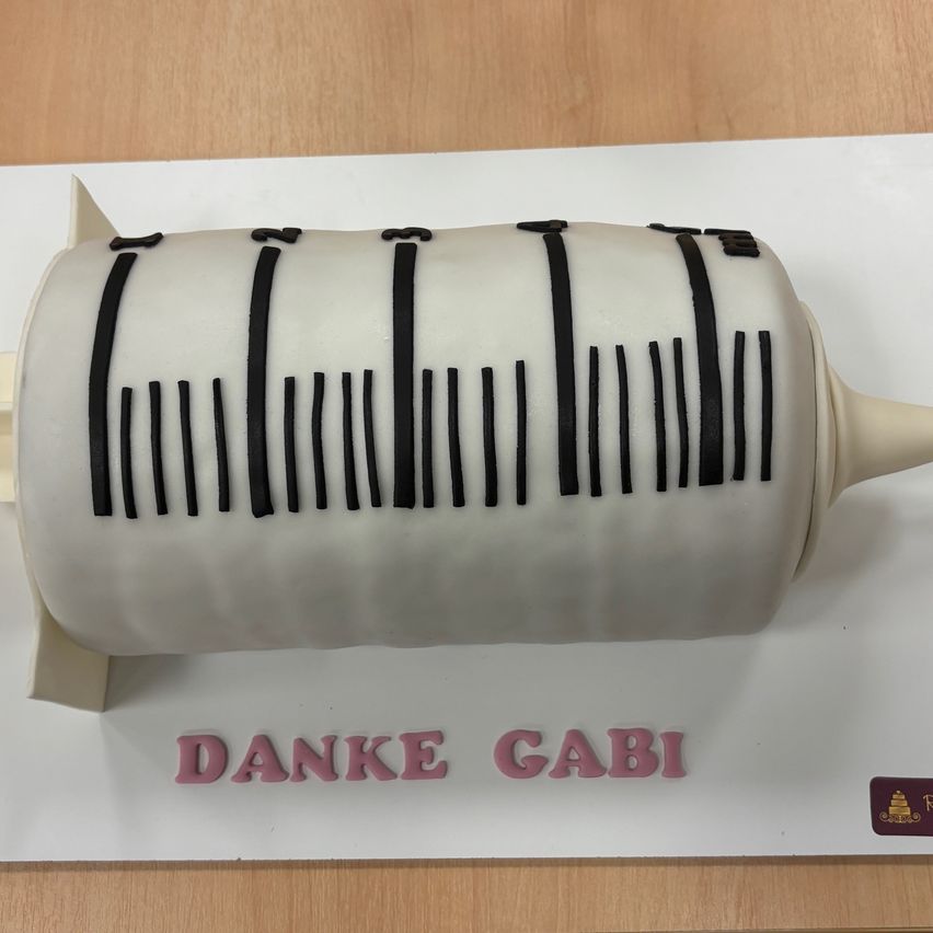 Ein Kuchen in Form einer Spritze mit einem Barcode-Muster, mit der Aufschrift 'Danke Gabi' darunter. Der Kuchen steht auf einer weißen Oberfläche, wahrscheinlich einem Kuchenkarton.