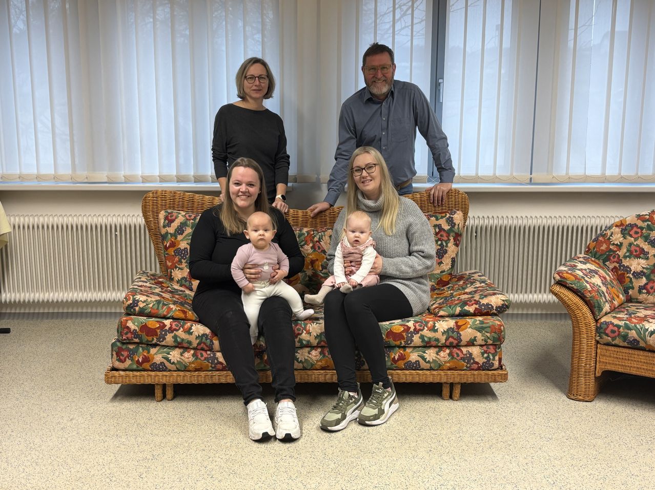 Eine Familie mit fünf Personen, darunter zwei Babys, sitzt auf einer blumenbedruckten Couch in einem Raum mit weißen Vorhängen. Die Erwachsenen stehen hinter der Couch.