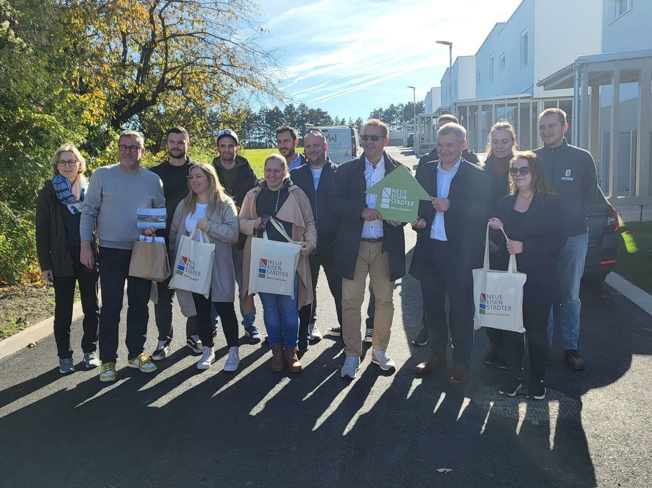 Eine Gruppe von Menschen steht vor einem Gebäude und hält weiße Taschen. Eine Person hält ein grünes Schild mit weißem Text und eine andere eine Papier.