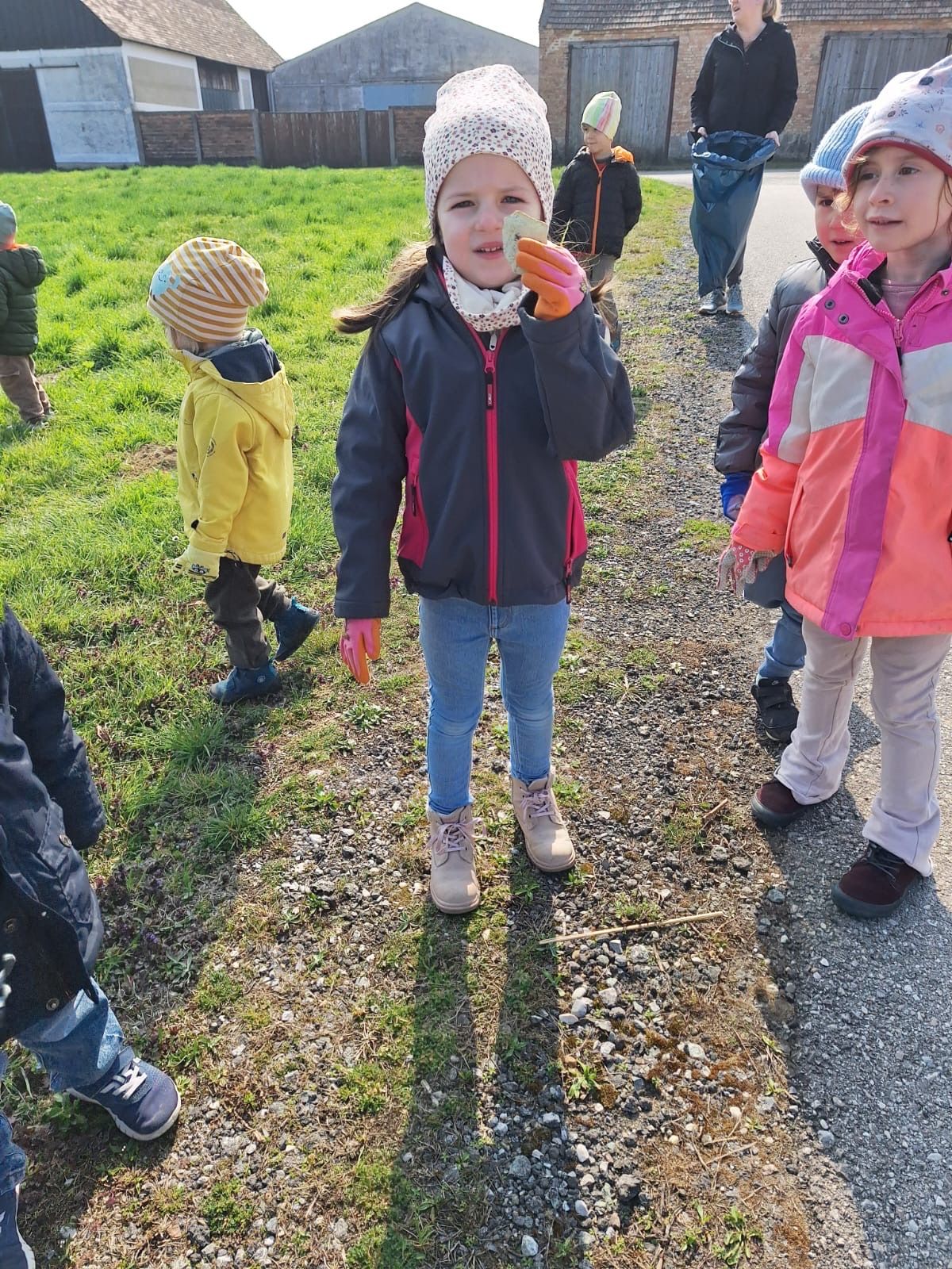A group of children walk along a gravel path. A girl in the center holds a piece of food. Behind her, a child in a yellow jacket and another in a pink jacket are walking.