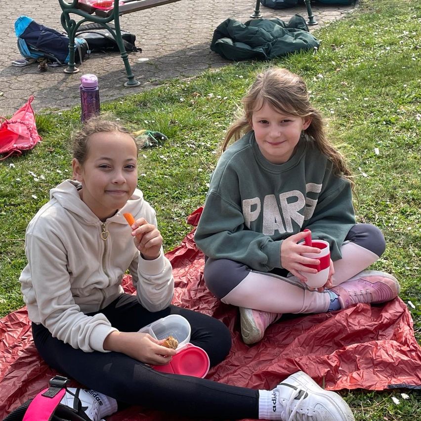 Two girls are sitting on a blanket in the park, smiling and enjoying a snack. One girl holds a carrot, and the other holds a cup. Bags and a bench are nearby.