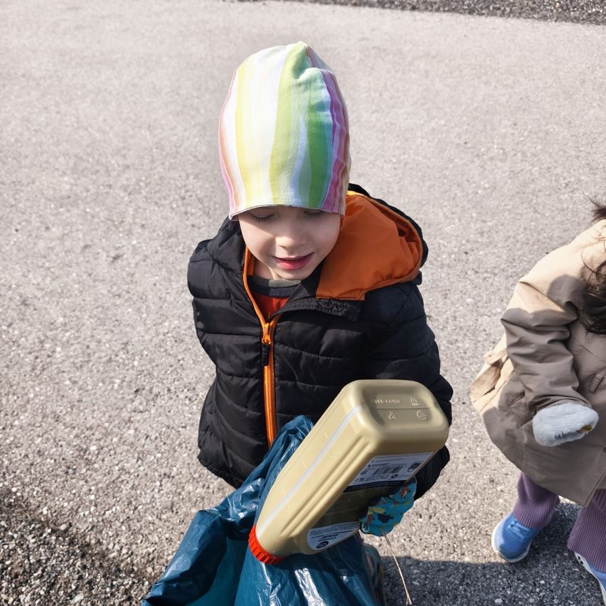 A child in a colorful hat and a black jacket holds a container of oil in a plastic bag, standing on a gravel road. Another child stands behind.