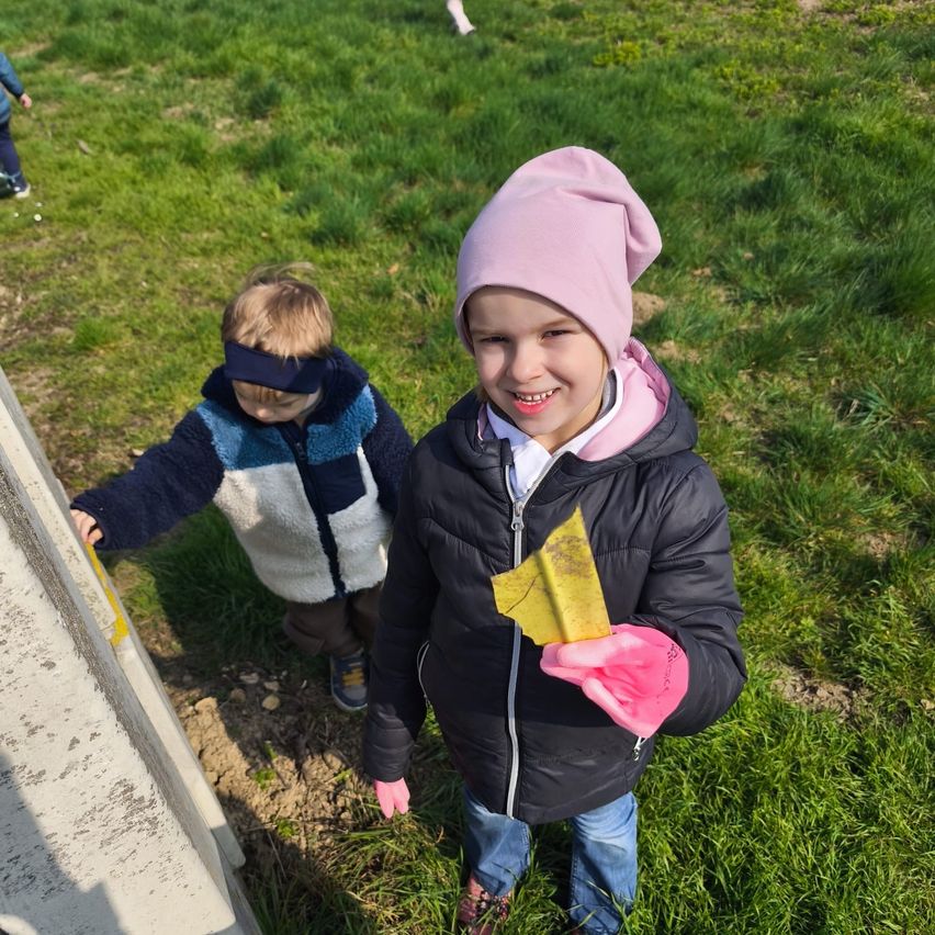 Two children walk on grass. The girl holds a yellow leaf, and the boy wears a blindfold. Another child is in the background.