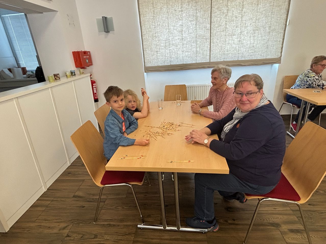 Three adults and two children sit around a table with straws, perhaps playing a game. A woman smiles, and another woman looks thoughtful.