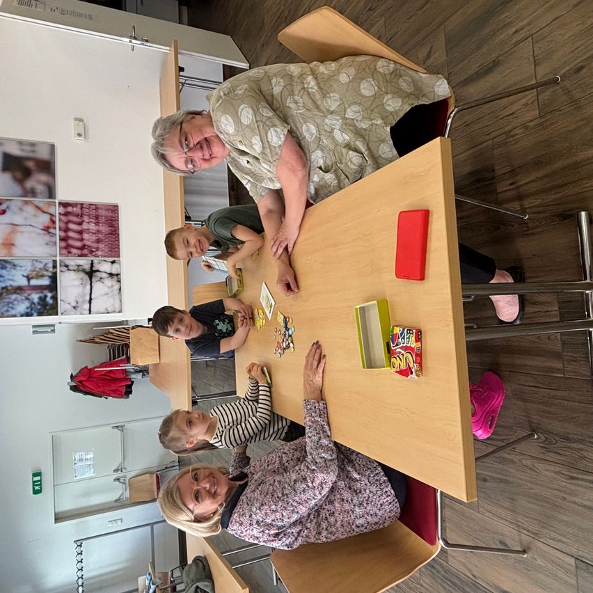 A group of people, including a woman and children, are sitting around a wooden table playing a board game. The woman is smiling.