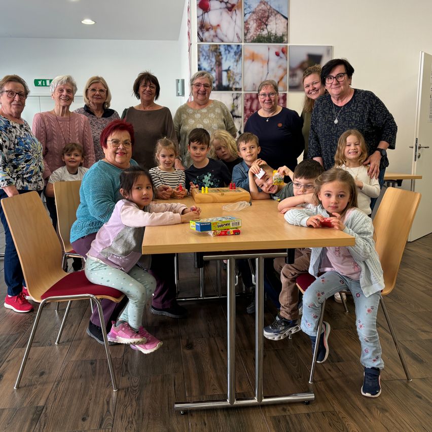 A group photo shows adults and children seated around a table, with toys and snacks visible. Some adults are standing behind the children, smiling and looking at the camera. The setting appears to be a community gathering.