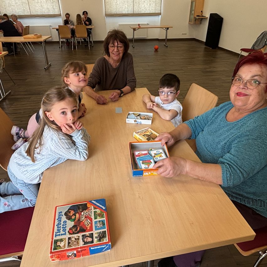 A group of people, including a young girl, boy, and elderly women, are sitting around a table playing a card game. The woman is holding a box with cards. In the background, a woman is sitting at a table with other people.