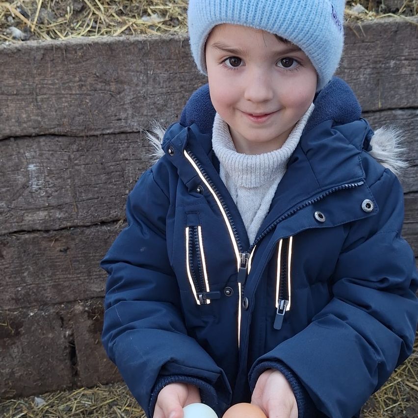 A smiling young child in a blue jacket and beanie holds two eggs, standing outside near a wooden wall.