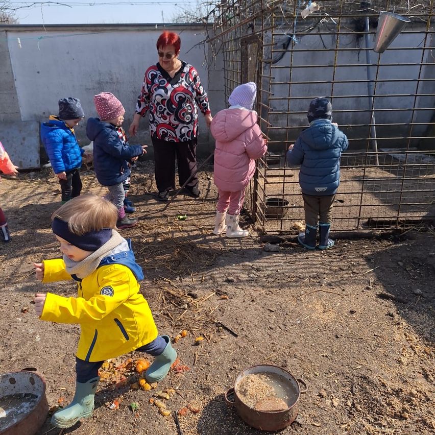 An older woman with red hair stands near a metal cage, with several children in winter clothing standing outside it. One child in a yellow jacket and green boots is looking down at something.
