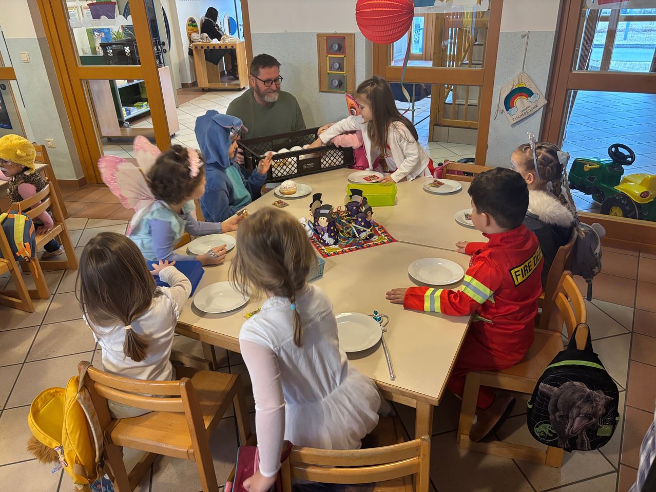 Eine Gruppe von Kindern in Kostümen sitzt um einen Tisch herum mit Tellern und Essen. Ein erwachsener Mann steht hinter ihnen und beaufsichtigt sie möglicherweise. Der Tisch ist mit einem bunten Tuch bedeckt und hat Spielzeug darauf. Im Hintergrund befindet sich eine Tür.