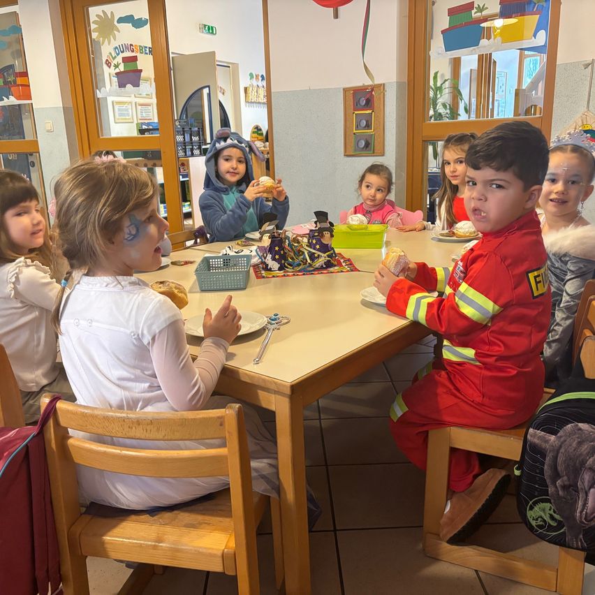Eine Gruppe von Kindern in einem Klassenzimmer sitzt am Tisch und genießt ihr Essen. Ein Kind trägt ein Feuerwehrmann-Kostüm.