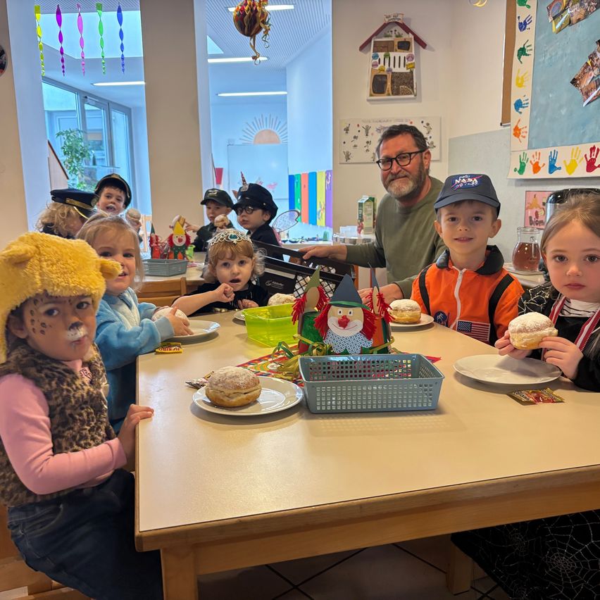 Eine Gruppe von Kindern in Kostümen, darunter ein Löwe und eine Hexe, sitzen um einen Tisch mit Essen, während ein erwachsener Mann hinter ihnen steht.