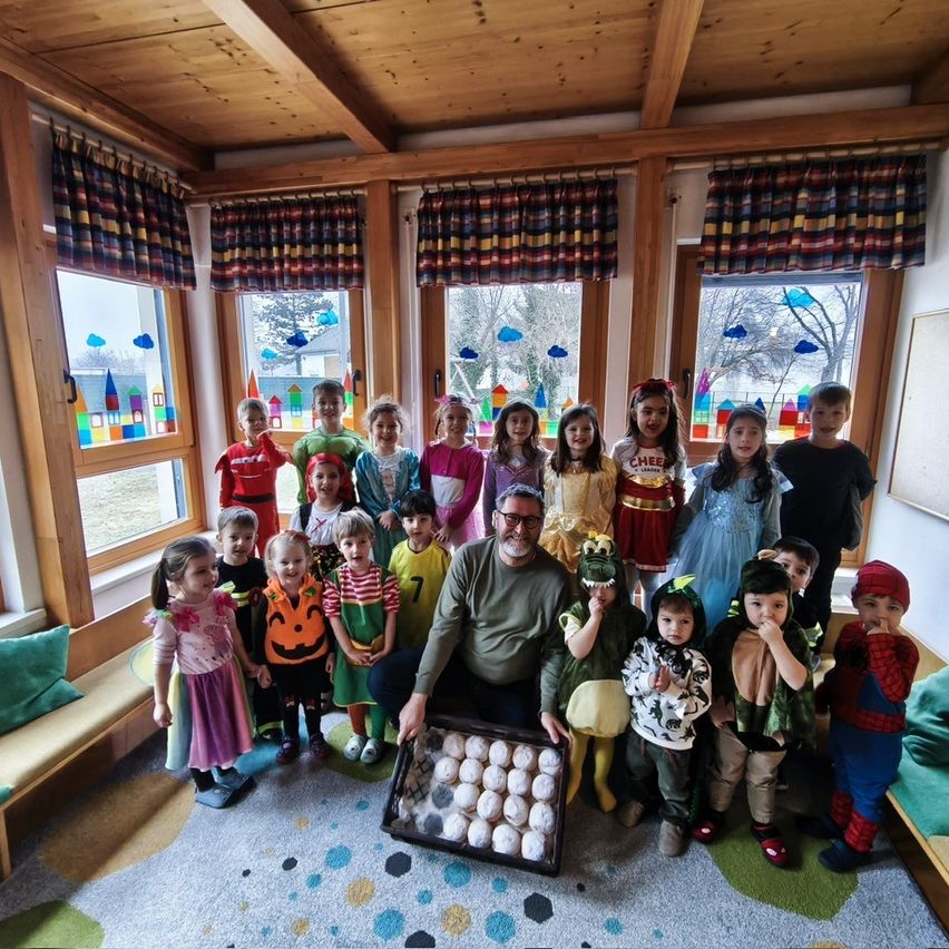 Eine Gruppe von Kindern und einem Erwachsenen posiert für ein Foto in einem Klassenzimmer. Der Erwachsene hält eine Tablett mit Essen. Die Kinder tragen Kostüme und stehen hinter dem Erwachsenen. Der Raum hat große Fenster und Holzbalken an der Decke.
