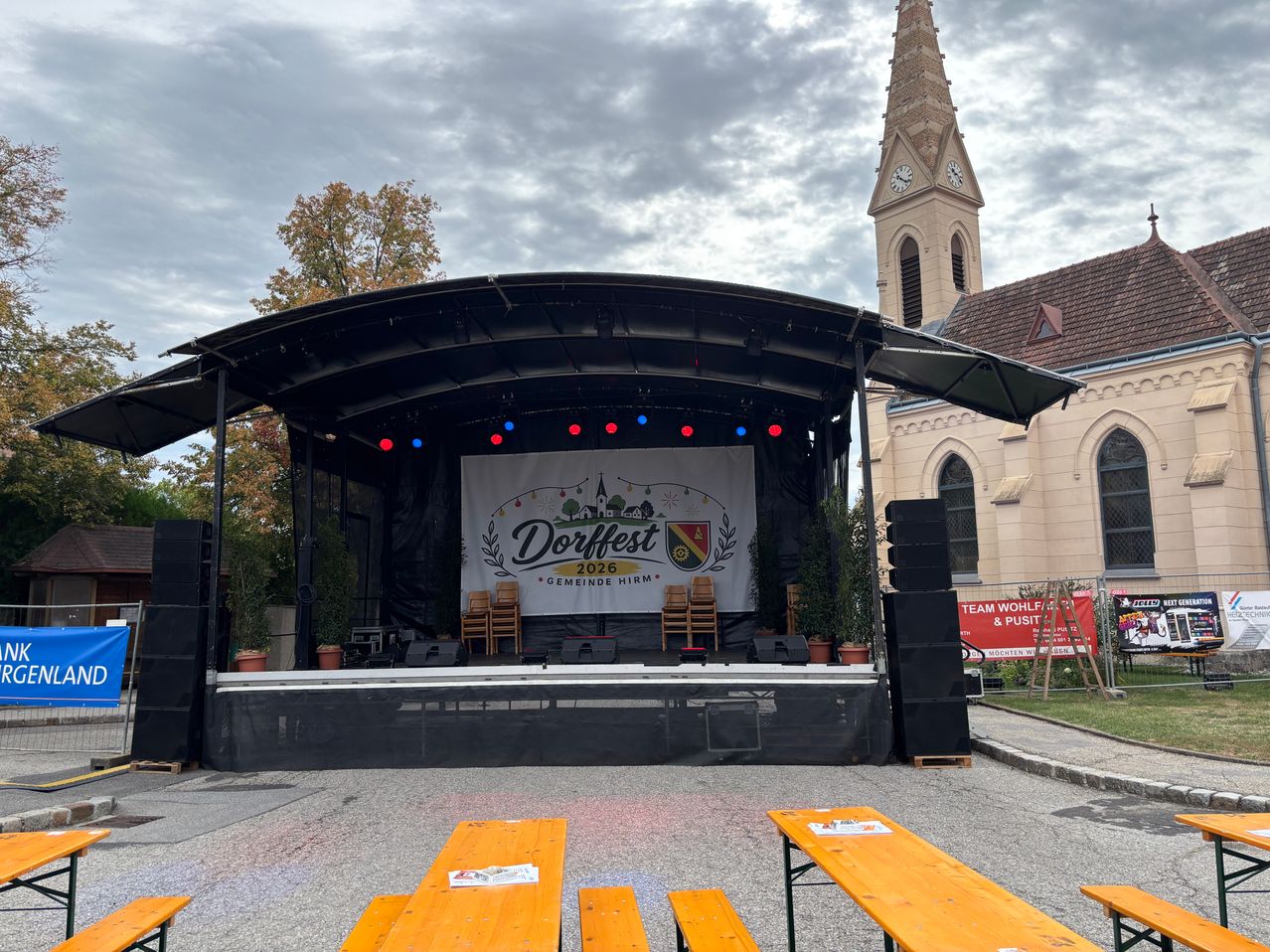 Eine Bühnenaufstellung für Dorffest 2026 steht vor einer Kirche mit bewölktem Himmel. Die Bühne hat ein Banner, Stühle und Lautsprecher. Davor stehen Tische und Bänke.