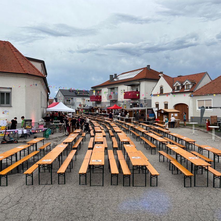 Eine Straßenszene mit in Reihen angeordneten Holzbänken, einem weißen Zelt und Gebäuden mit Flaggen im Hintergrund.