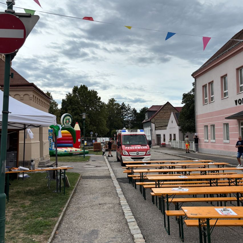 Eine Straßenszene mit einem Feuerwehrauto, Tischen und einem Hüpfburg. Der Himmel ist bewölkt, und es gibt Gebäude und Bäume im Hintergrund.