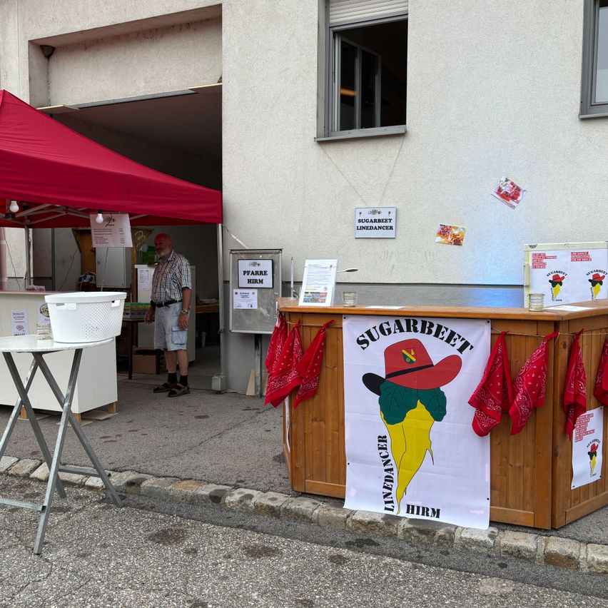 Ein Mann steht vor einem Gebäude mit einem roten Zelt. Ein Holztresen hat ein Banner mit der Aufschrift 'Sugarbeet Linedancer'.