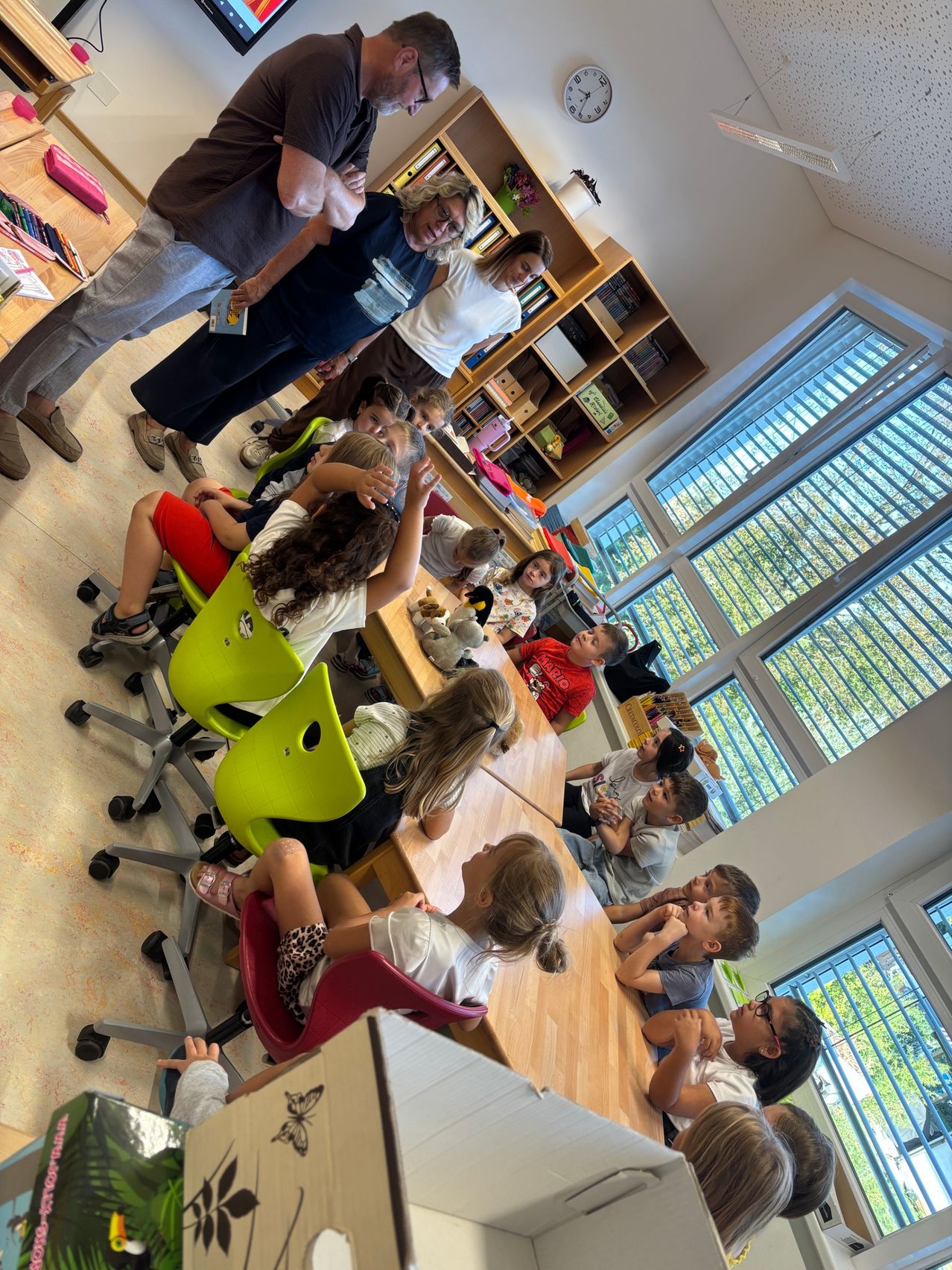 Eine Gruppe von Kindern sitzt aufmerksam um einen Tisch in einem Klassenzimmer und hört einer Erwachsenen zu, die ein Buch in der Hand hält. Der Raum ist gut beleuchtet mit großen Fenstern und Regalen voller Bücher.
